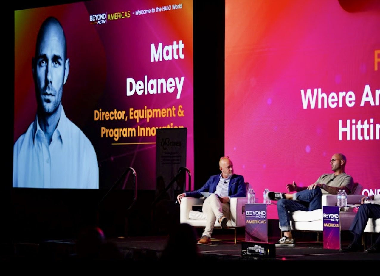 Matt Delaney seated on stage during a panel discussion, his name and title shown on the screen behind him