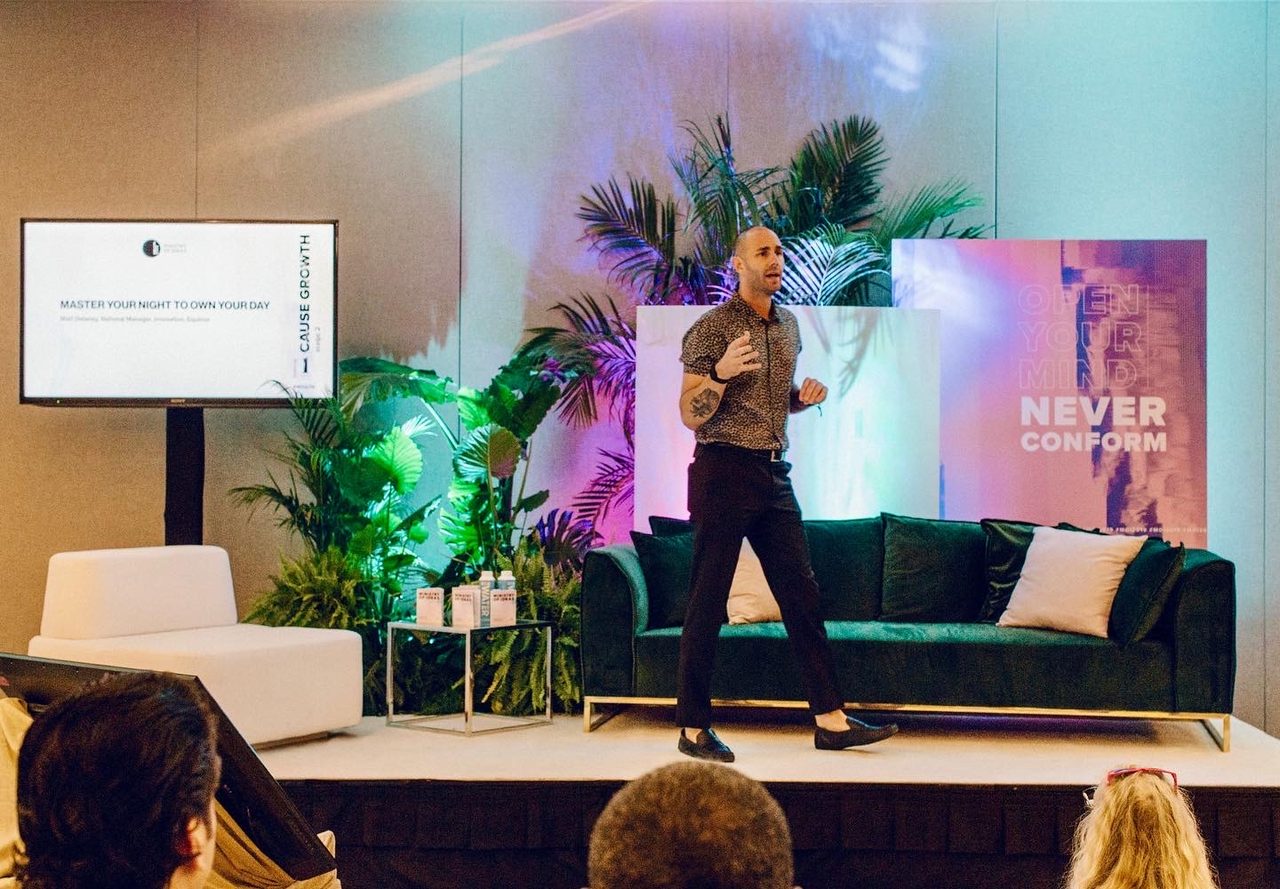Matt Delaney presenting to an audience on a stage set with plants and a lounge sofa